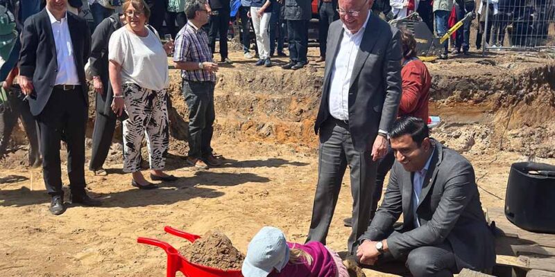 Dr. Wjahat Waraich hilft Kindern beim Spatenstich in der Baugrube, dahinter die Gäste des Baueröffnungsfestes.