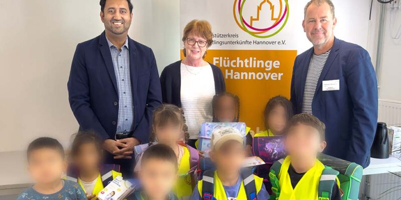 Dr. Wjahat Waraich übergibt mit zwei Helfenden Schulranzen an Kinder in der Flüchtlingsunterkunft; die Gesichter der Kinder sind unkenntlich gemacht.