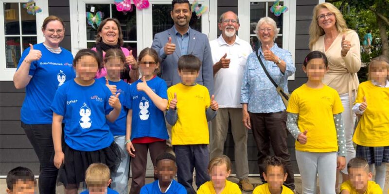 Dr. Wjahat Waraich steht mit Kindern in blauen und gelben T-Shirts vor dem neuen Gartenhaus der Grundschule Bothfeld.